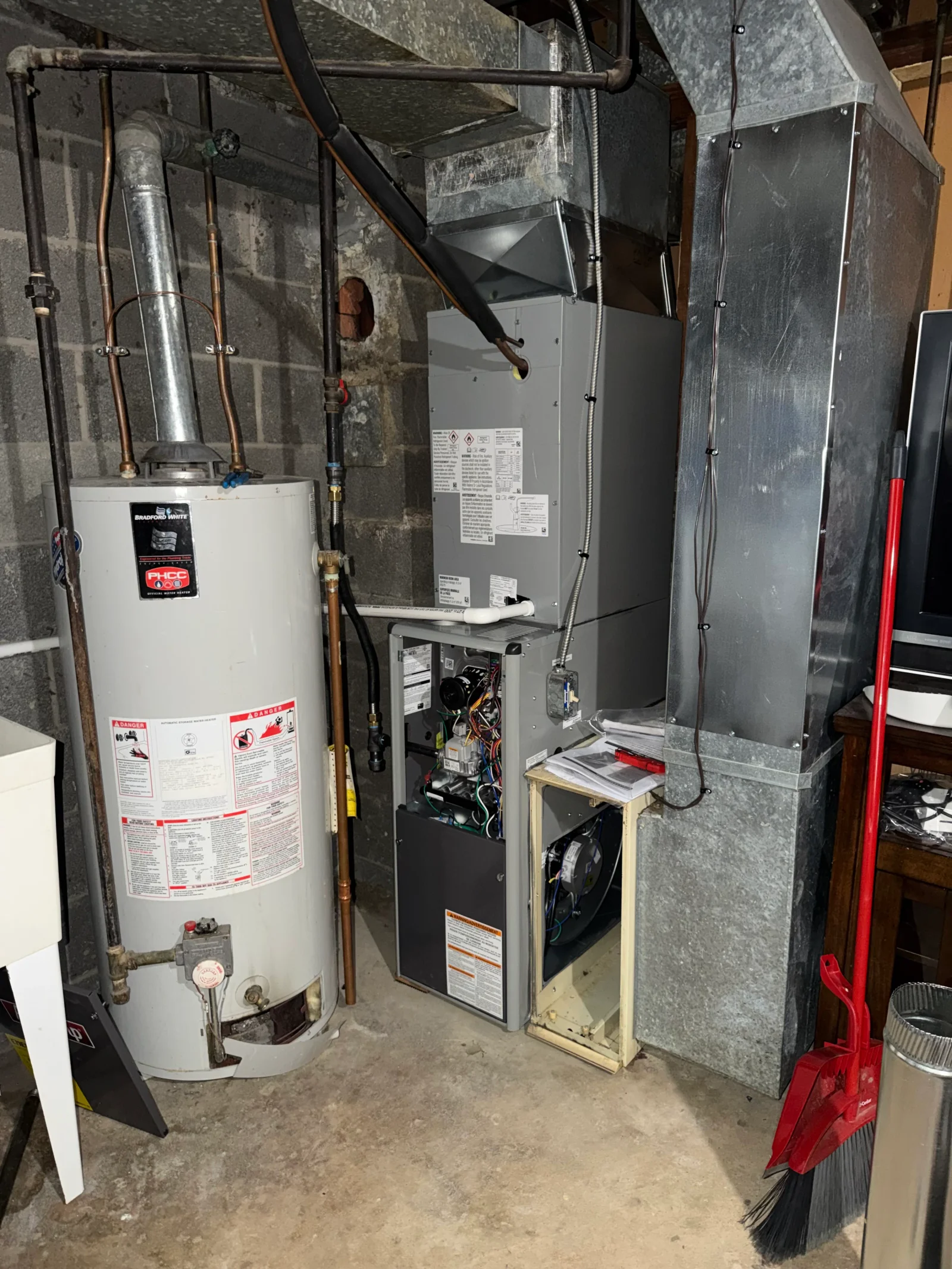 Basement gas furnace with open access panel beside Bradford White gas water heater, galvanized metal ductwork and flue pipe