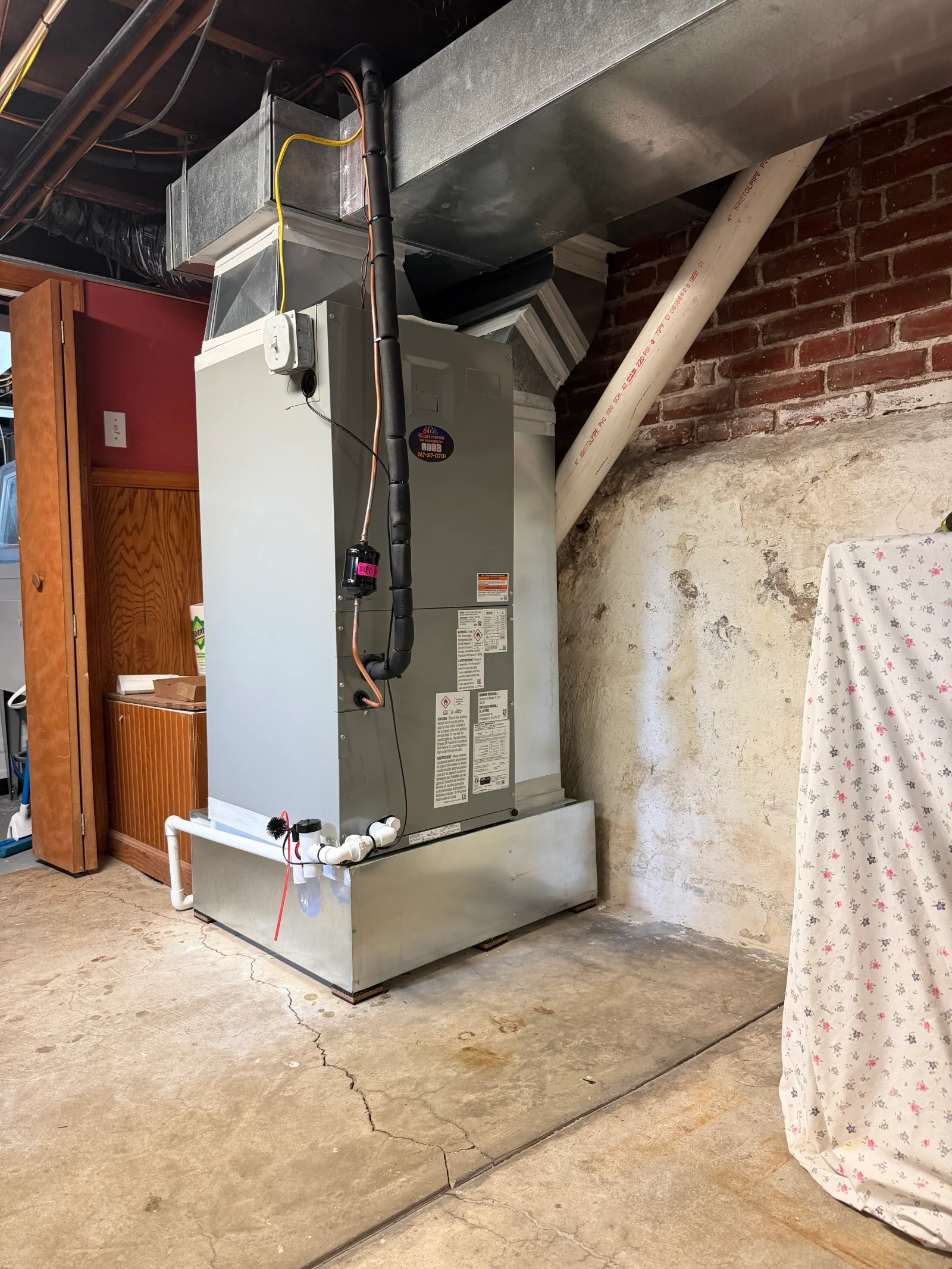 Gray air handler on steel platform with galvanized ductwork, insulated copper refrigerant lines, and PVC condensate drain piping in a basement