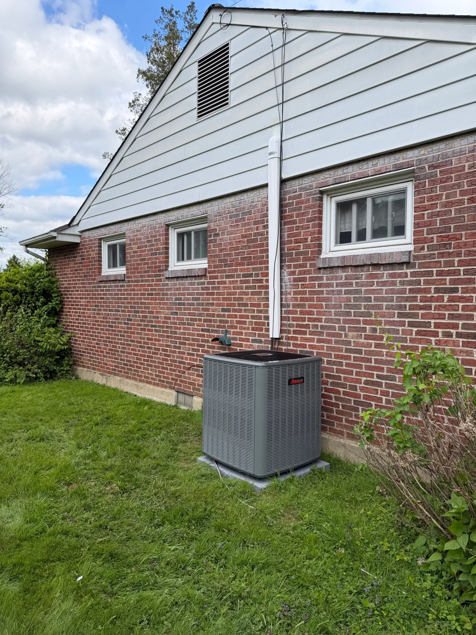 Gray Amana outdoor AC condenser on concrete pad beside red brick home with white vinyl siding