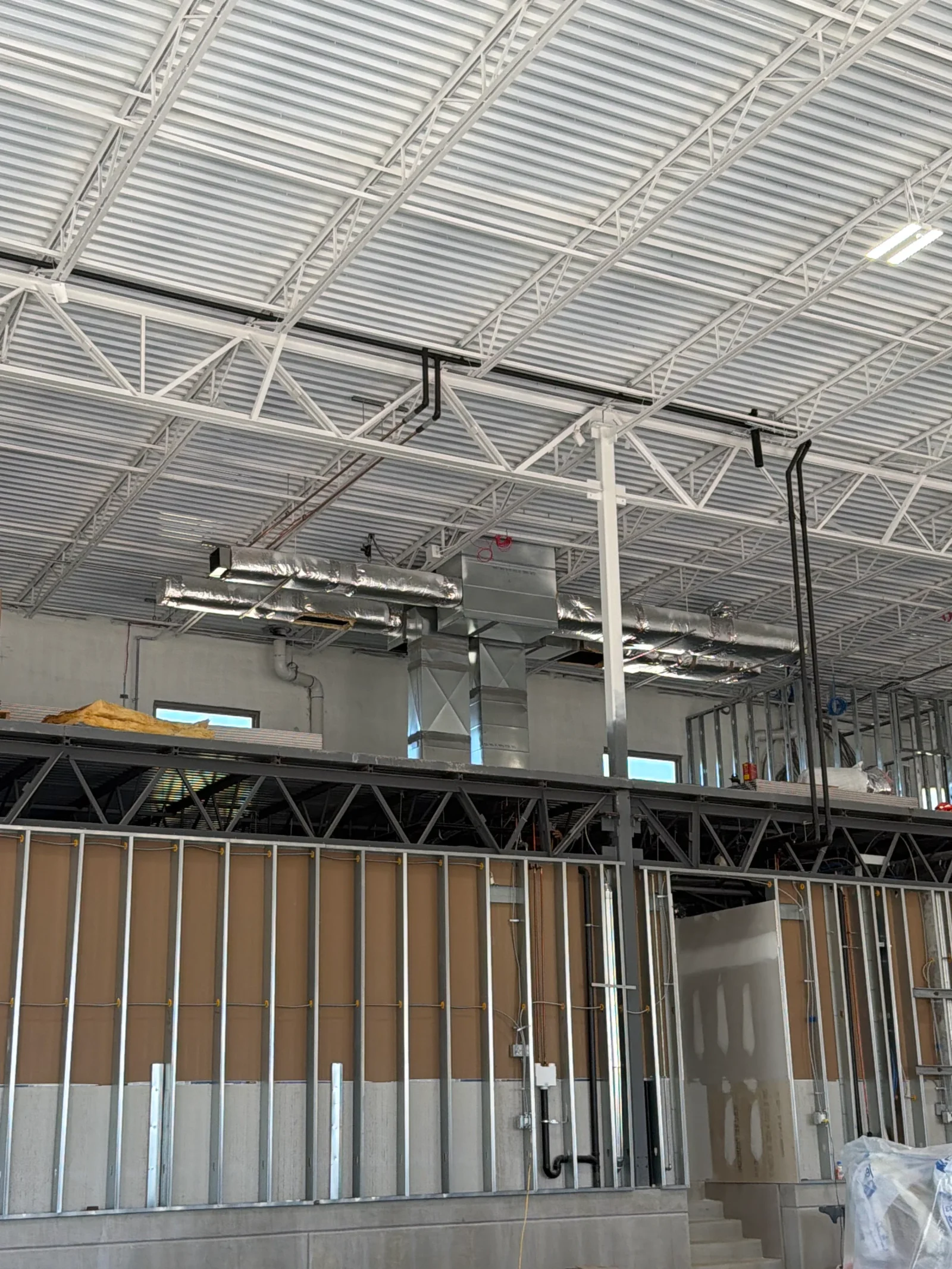 Insulated silver flex and galvanized sheet metal ductwork suspended from white steel roof trusses in a commercial building under construction