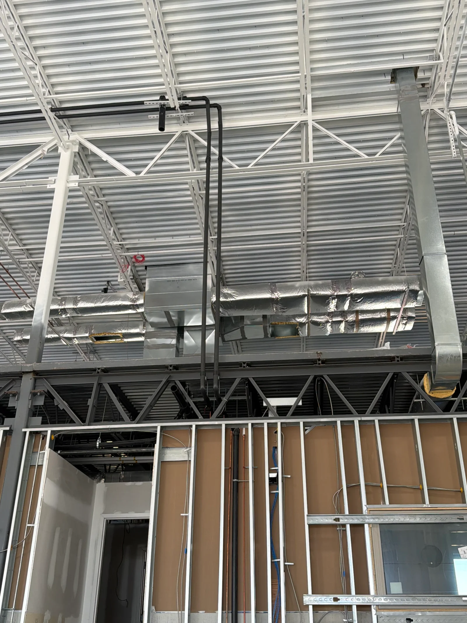 Overhead commercial HVAC ductwork with foil-wrapped insulation, galvanized sheet metal, and black piping under steel truss framing