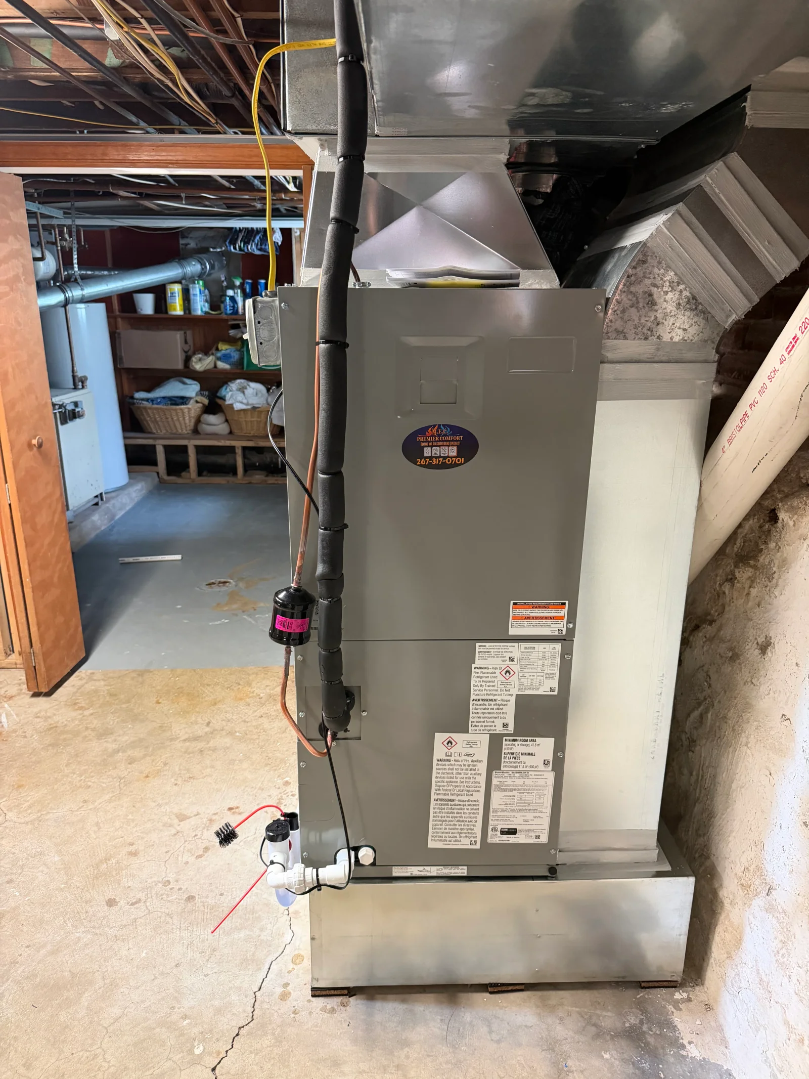 Gray air handler with insulated copper refrigerant line, white PVC condensate drain, and sheet metal plenum in basement