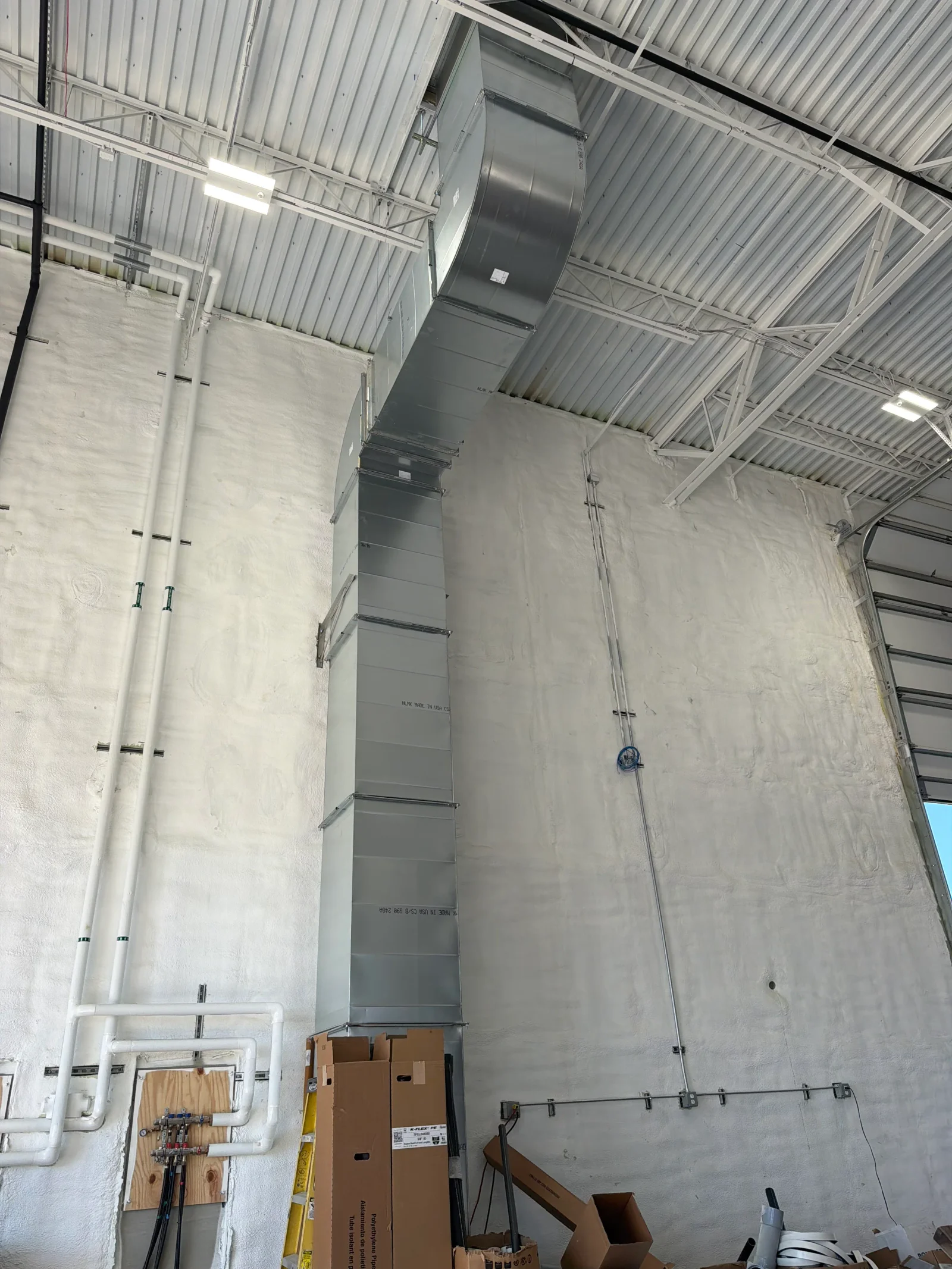 Galvanized sheet metal supply ductwork and PVC piping routed along spray-insulated walls in an industrial building