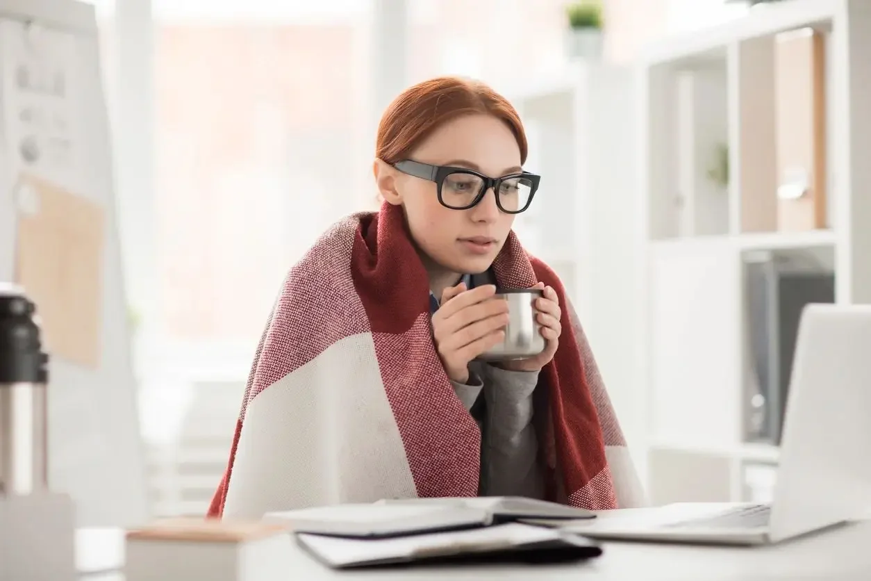 Woman in glasses wrapped in plaid blanket holding a stainless steel mug at a white desk with laptop and notebooks