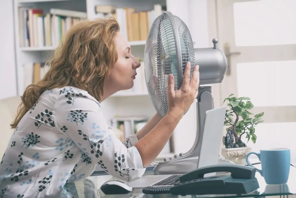 Woman feeling airflow from a silver desk fan at a glass desk with laptop and telephone