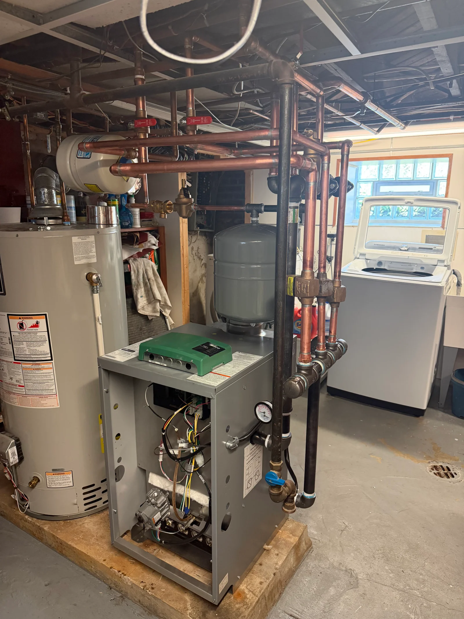 Gray boiler with green control module, expansion tank, and copper and black iron piping in a basement